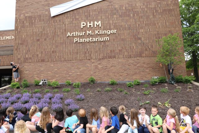 Children gathered in front of a brick building with a sign that reads 'PHM' and the name 'Arthur M. Klinger Planetarium'.