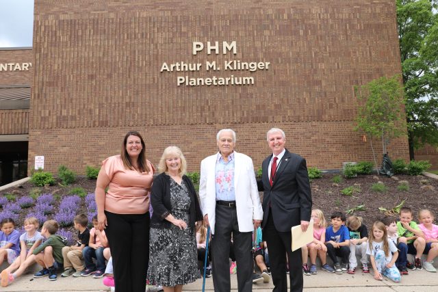 An elderly couple posing in front of a brick building with a sign that reads "PHM".