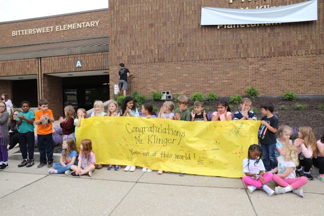 Group of children celebrating with a yellow banner, posing in front of a school building.