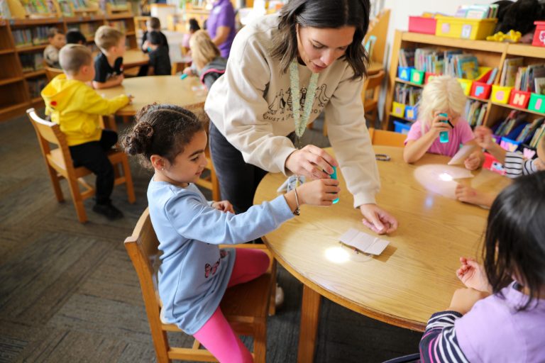 Woman in white shirt pouring blue liquid into cups at a table surrounded by children.