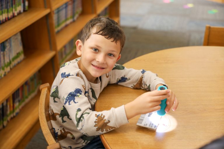 Young boy sitting at a table in a library, holding a flashlight.
