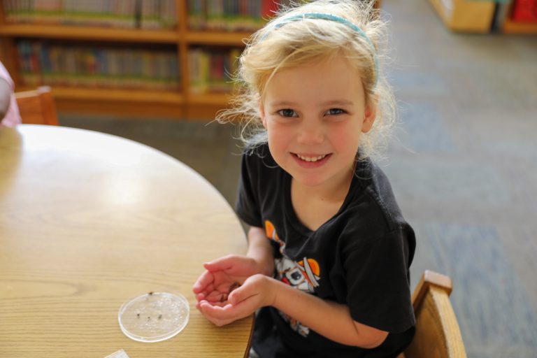 Young girl sitting at table in library, smiling at camera, holding a small object in her hand.