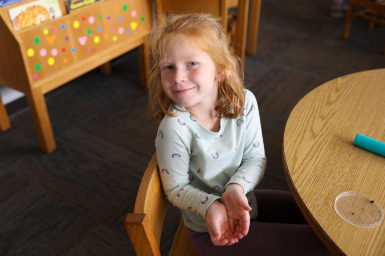 Little girl sitting at table in classroom, smiling at camera.