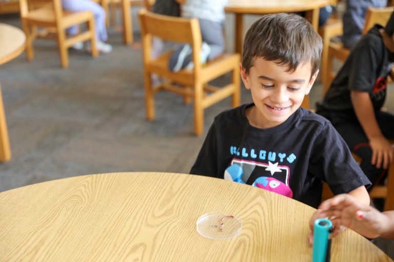 Young boy sitting at a table in a classroom setting, smiling and looking directly at the camera.