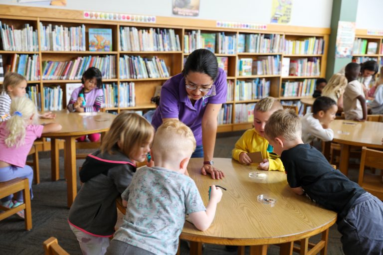 Woman standing behind table of children engaged in activity.