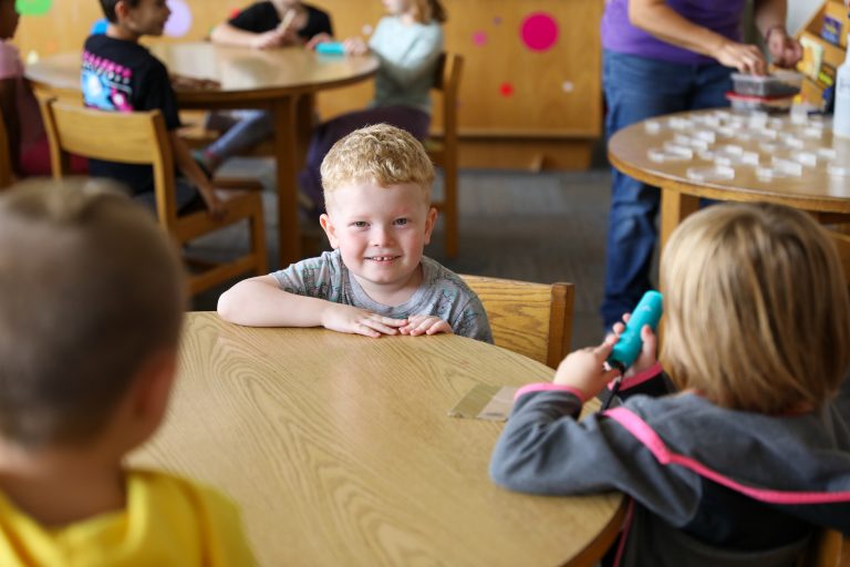 Small child sitting at table in school room, smiling at camera.