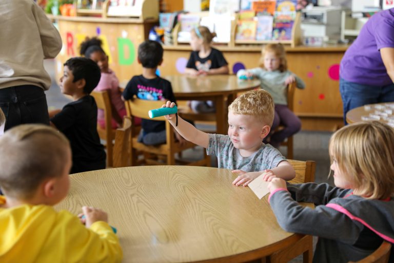 Children sitting at a table in a classroom. A young boy is holding a green toy. The teacher is standing behind the children.