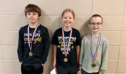 Three children posing in front of a wall with medals around their necks, each holding a medal, smiling and looking at the