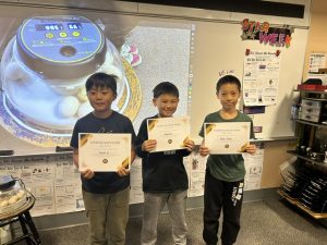 Three young boys proudly holding certificates in front of a classroom display.