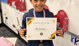 Young boy proudly holding a certificate of recognition in front of a bulletin board.